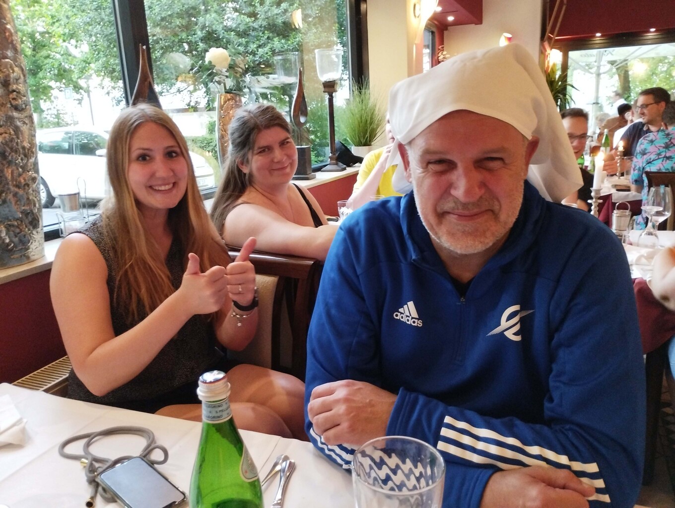 Mann mit einem Papierhut, der am Tisch sitzt und in die Kamera lächelt, neben ihm sitzt eine lachende Frau, die einen Daumen nach oben streckt