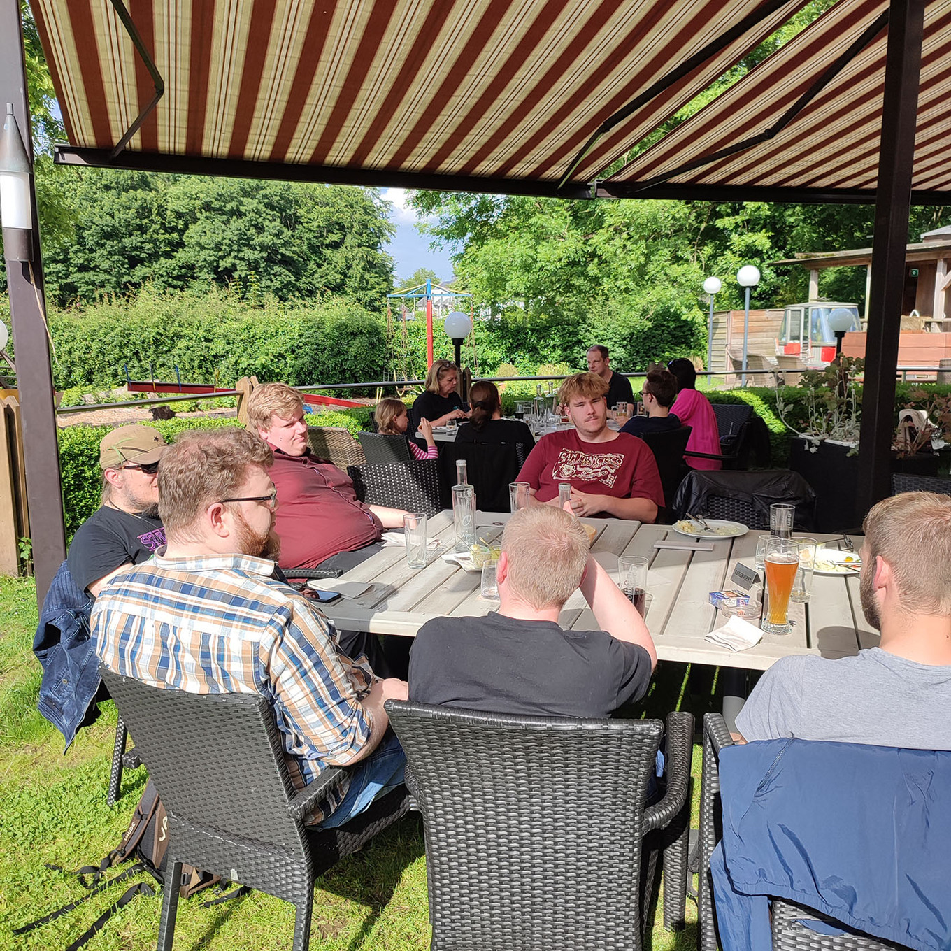 Gruppe von Menschen, die an einem sonnigen Tag im Freien unter einem gestreiften Sonnenschirm an einem Tisch sitzen und sich unterhalten 