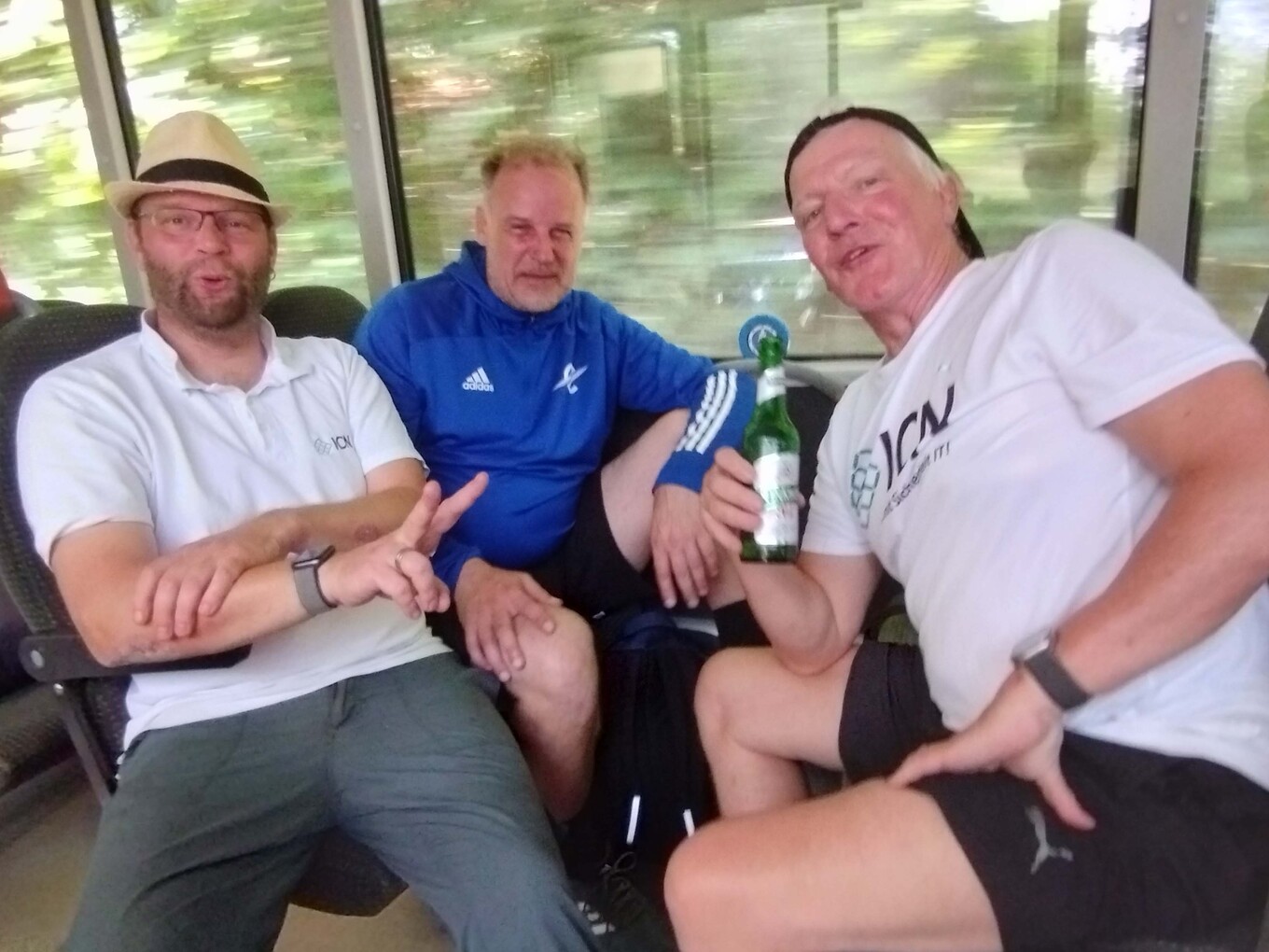 3 lächelnde Männer, die in einem Bus sitzen, einer der Männer trägt einen Hut und zeigt ein Friedenszeichen, ein anderer hält eine Flasche in der Hand 