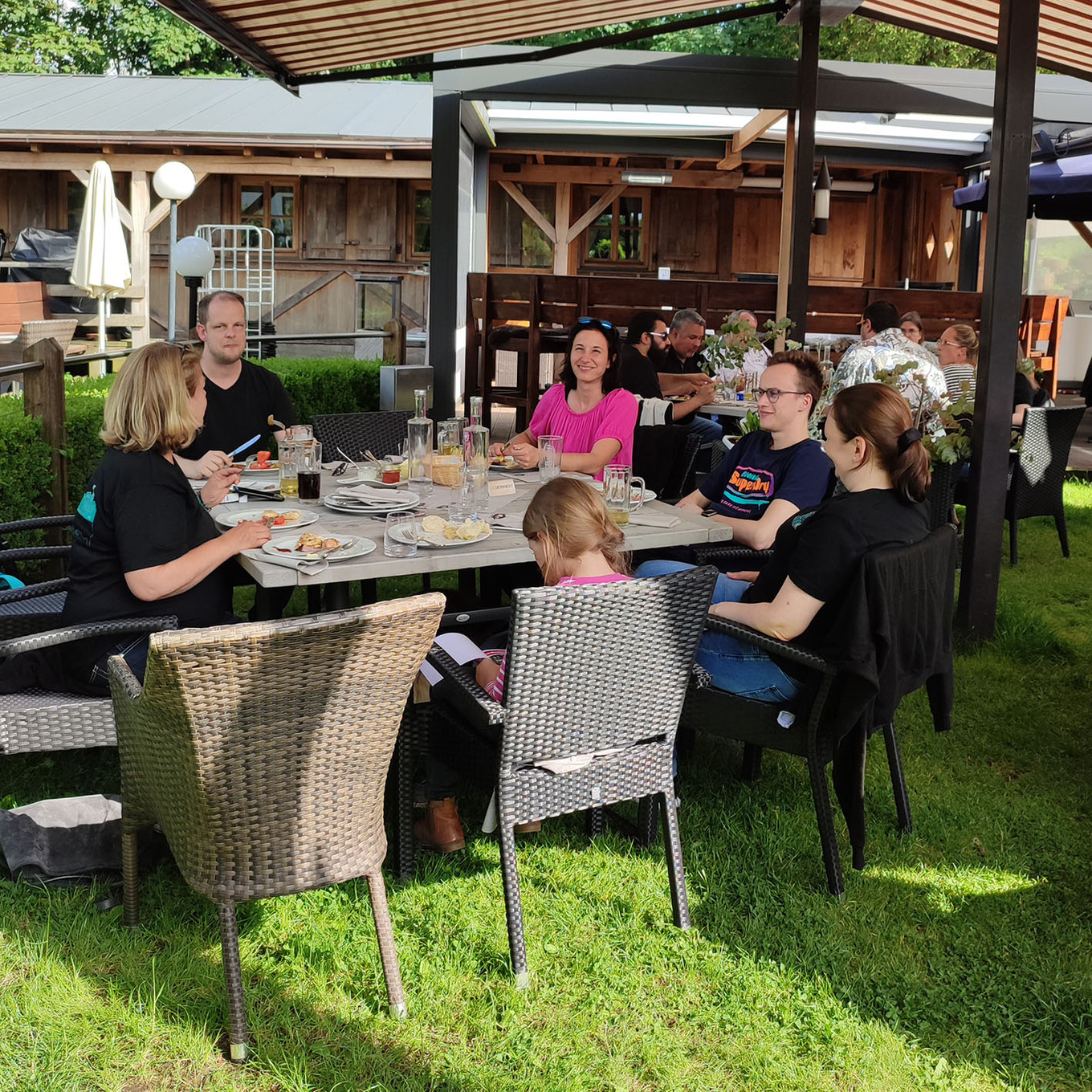 Gruppe von Erwachsenen und ein Kind, die gemeinsam an einem Tisch unter einem Sonnenschirm auf einer grünen Wiese im Freien essen 