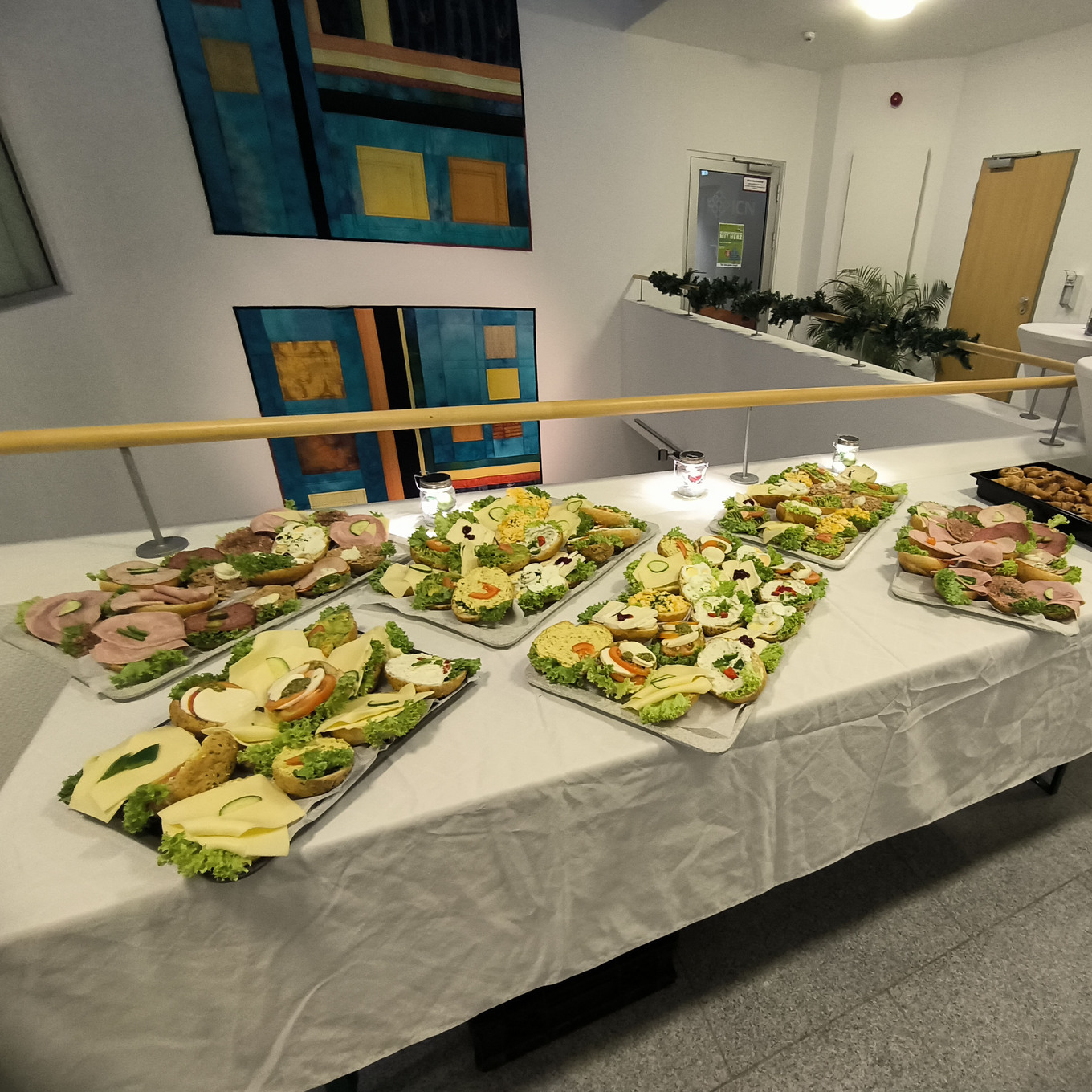 Buffet in einem Innenraum, auf einem langen Tisch, der mit einem weißen Tischtuch bedeckt ist, stehen mehrere Platten mit kleinen Sandwiches, Häppchen und Salaten