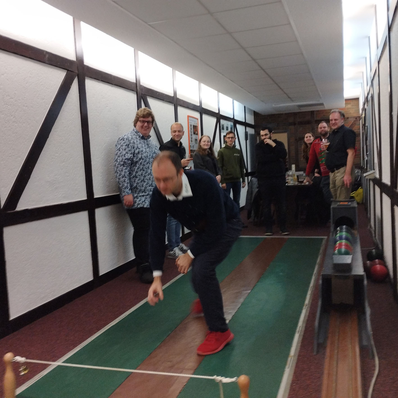 Mann in der Mitte, der sich über eine Kegelbahn beugt und gerade eine Kugel wirft, im Hintergrund steht eine Gruppe von Menschen und schaut zu