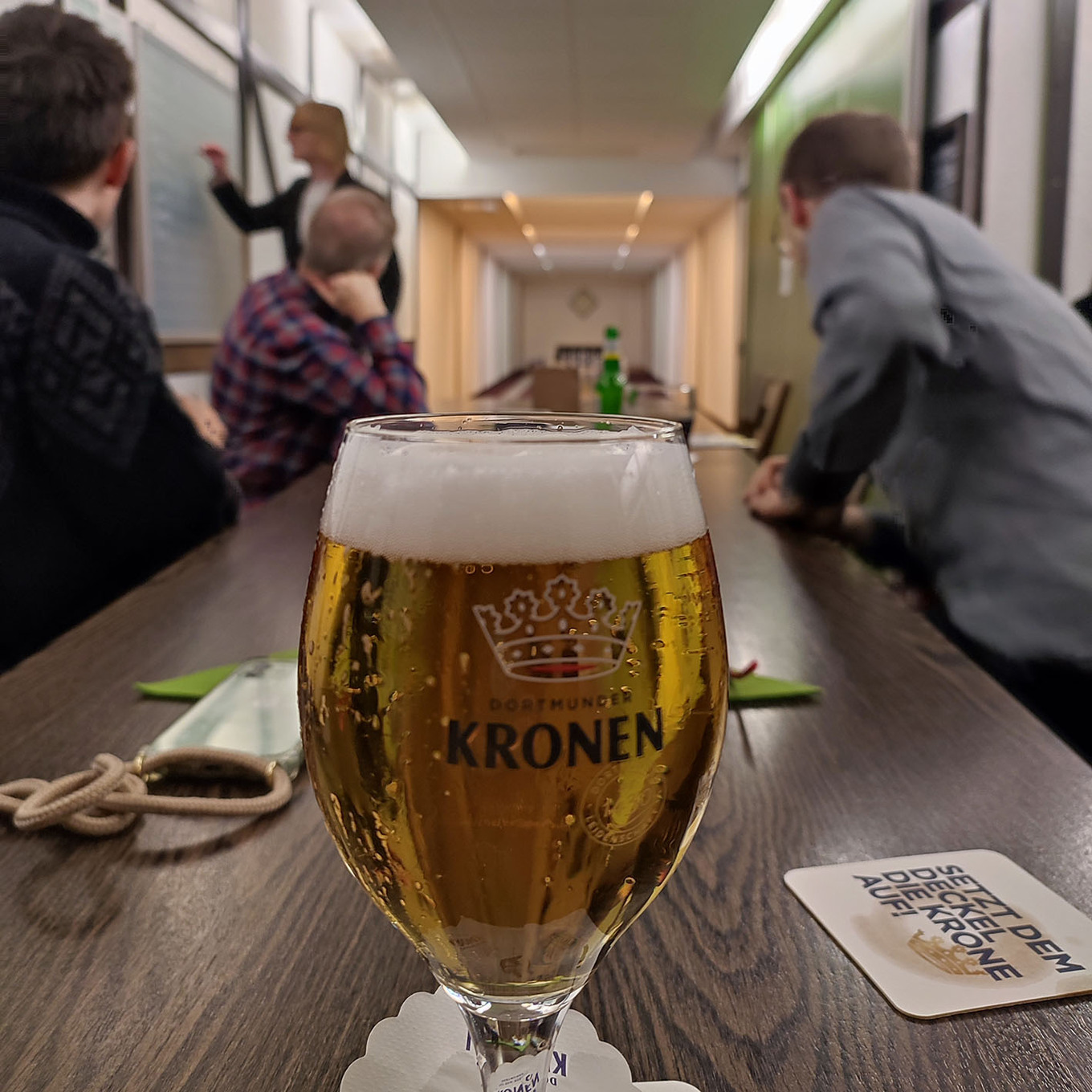 niedrige Perspektive auf ein Glas Bier mit einem Kronen-Logo, das auf einem Tisch in einem Konferenzraum steht, in dem im Hintergrund mehrere Personen sitzen 