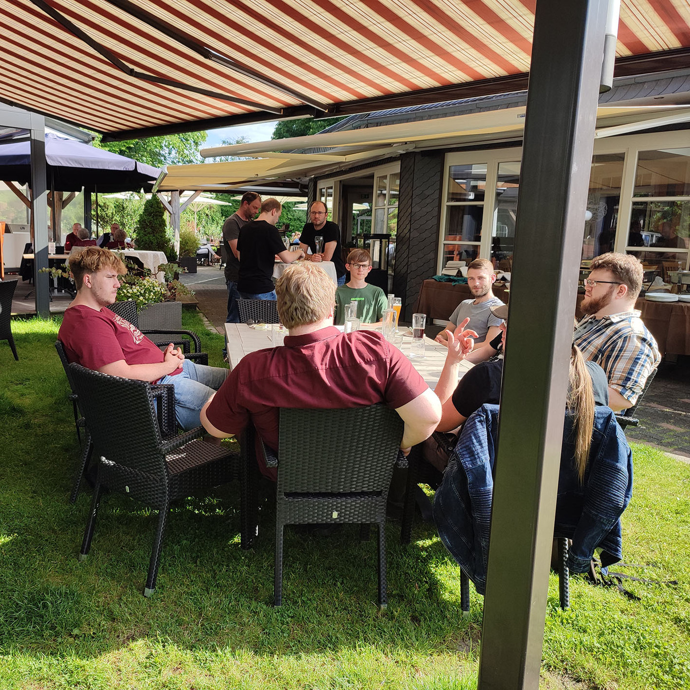 Gruppe von Menschen, die an einem sonnigen Tag unter einem gestreiften Sonnenschirm auf einer Wiese zusammensitzen 