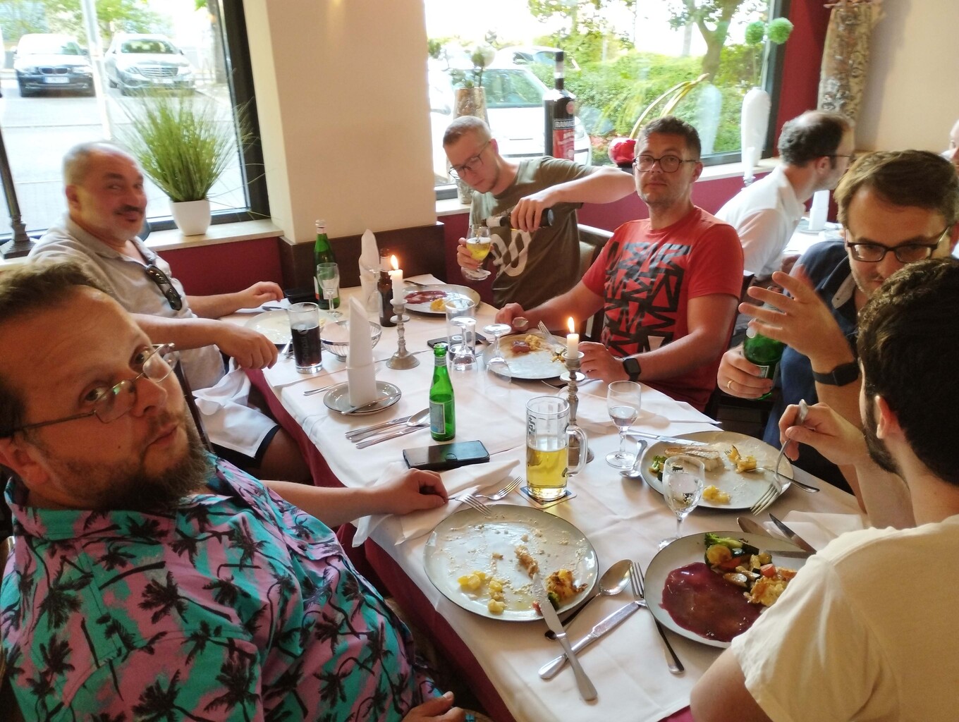 Gruppe von etwa 7 Männern, die in einem Restaurant an einem Esstisch sitzen, lachen und in die Kamera schauen, der Tisch ist mit Tellern, Gläsern und Flaschen gedeckt