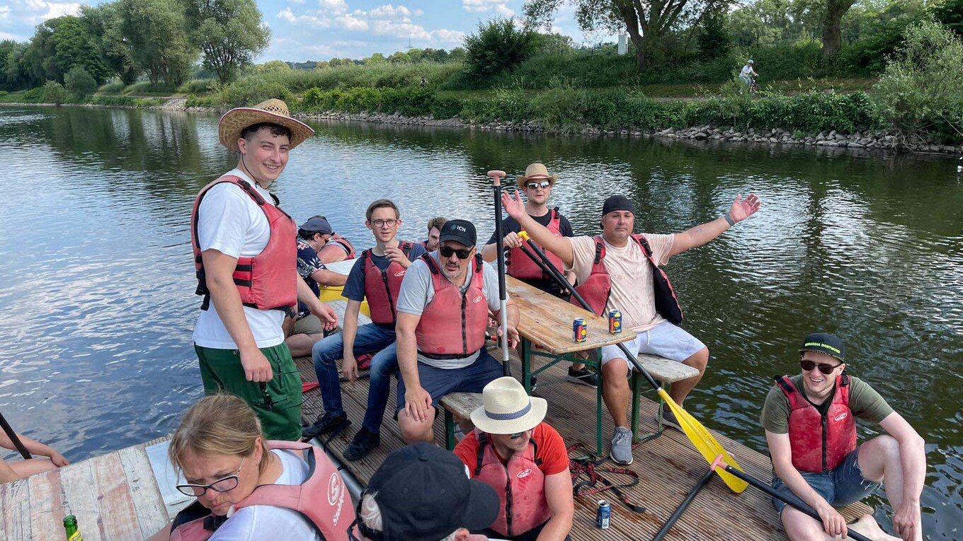 Gruppe von etwa acht Personen, die in Schwimmwesten und mit Hüten auf einem selbstgebauten Floß auf einem See sitzen und in die Kamera winken 