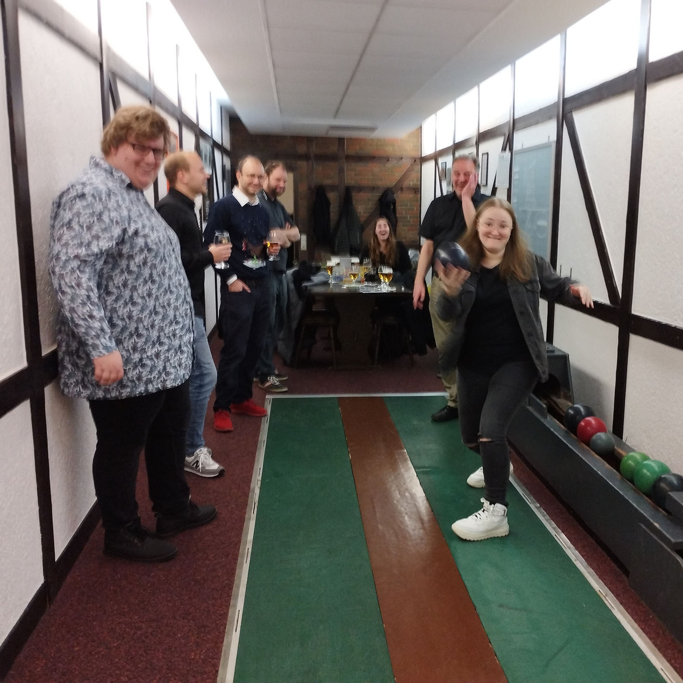 lachende Gruppe von etwa 8 Personen in einer Kegelbahn, eine junge Frau im Vordergrund hält gerade eine Kegelkugel in der Hand, um sie zu werfen