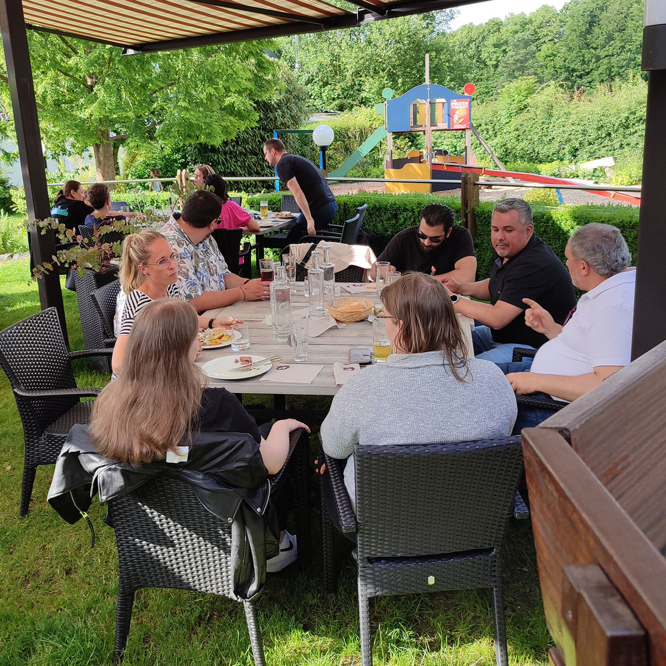 Gruppe von Menschen, die an einem sonnigen Tag im garten an einem großen Holztisch sitzen und sich unterhalten 