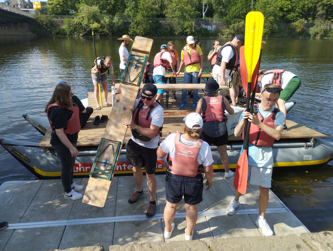 große Gruppe von Erwachsenen und Kindern auf einem selbstgebauten Floß auf einem See, von denen die meisten Schwimmwesten tragen und einige Paddel halten 