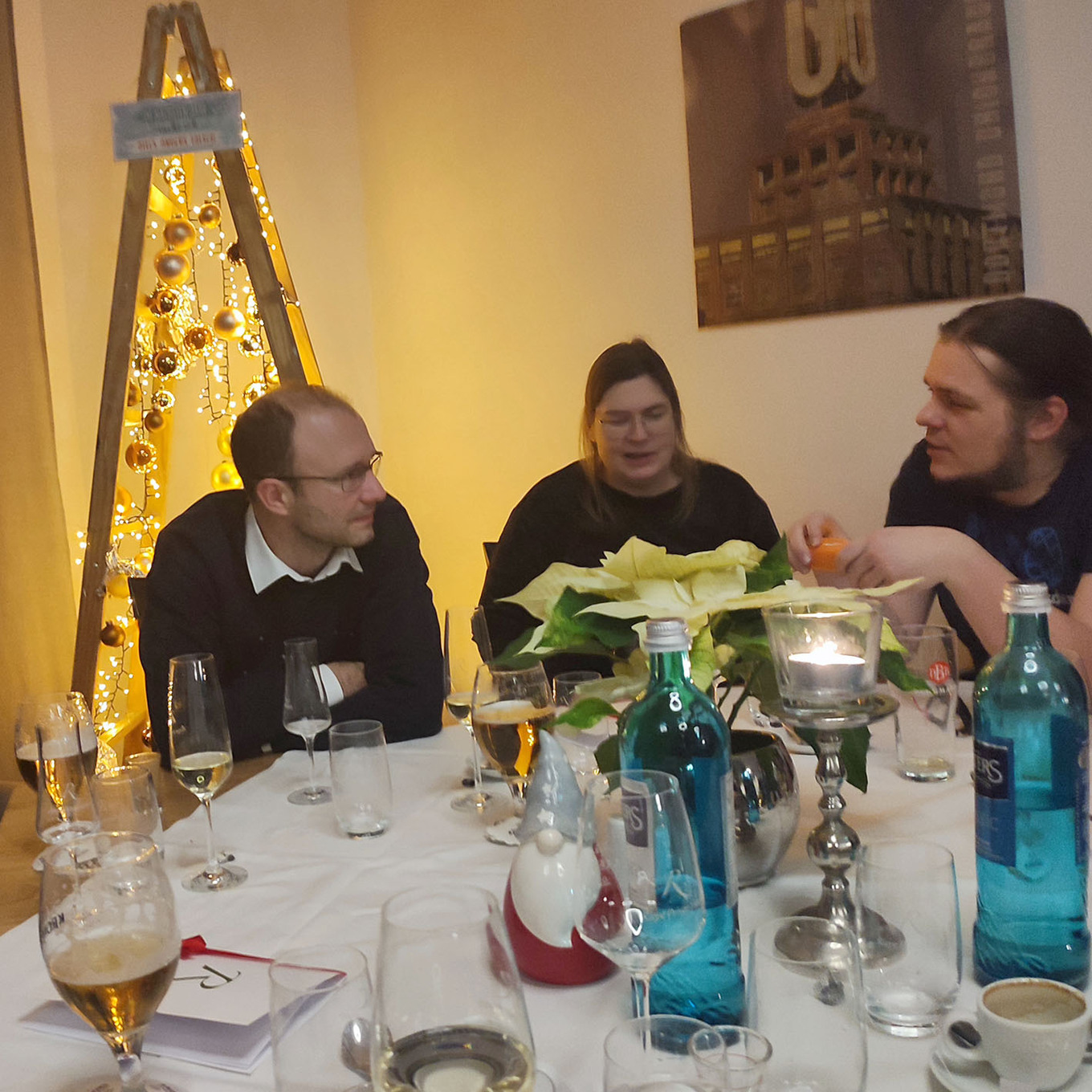3 Personen sitzen an einem festlich gedeckten Tisch und unterhalten sich, während im Hintergrund ein beleuchteter Weihnachtsbaum steht 