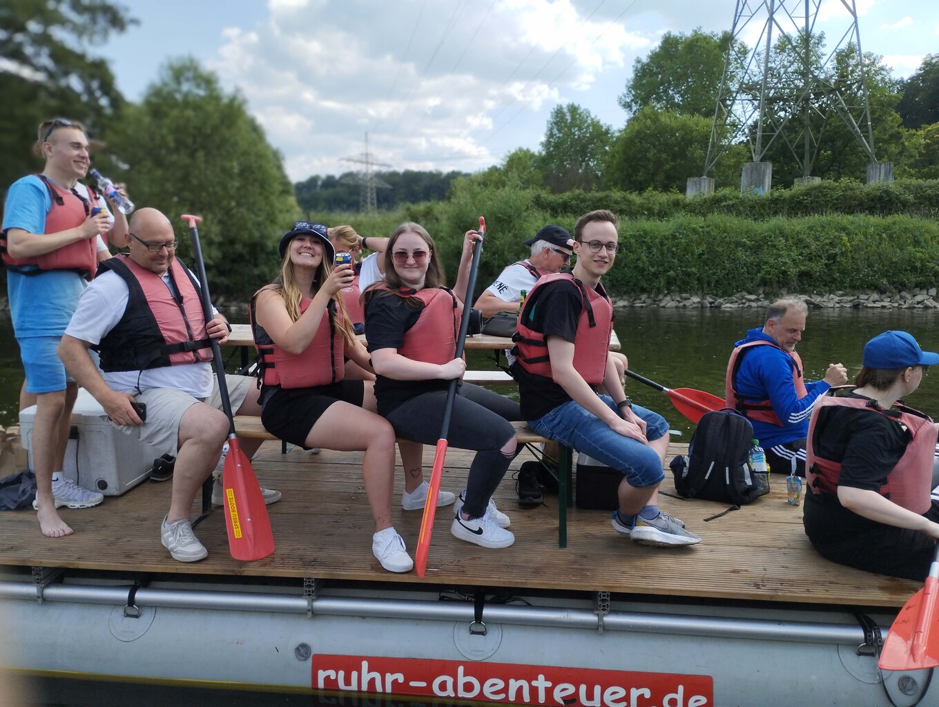Gruppe von 9 Personen in Rettungswesten, die auf einem Floß auf einem See sitzen, einige Personen haben Paddel in den Händen 