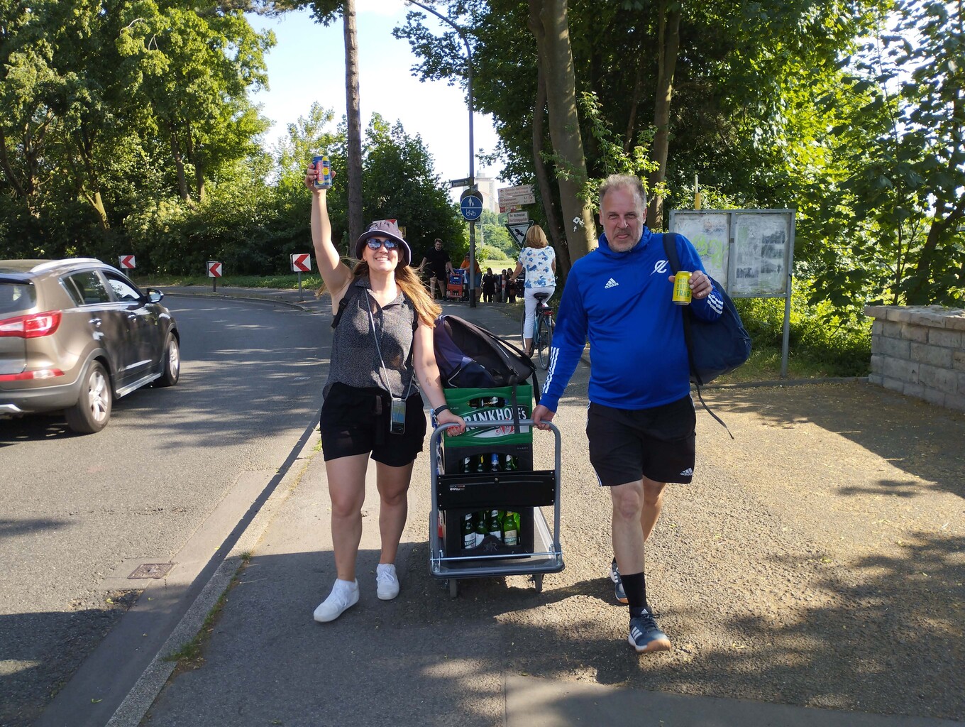 lachende Frau und Mann, die eine Sackkarre mit einer Getränkekiste schieben, die Frau hebt eine Flasche in der Hand hoch