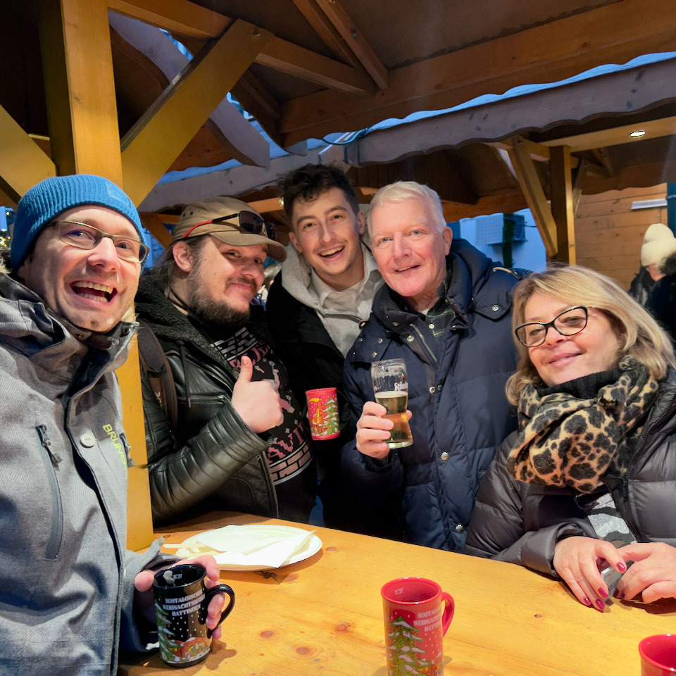 Gruppe von fünf Personen, die lachend mit Tassen und Bechern in den Händen an einem Stand auf einem Weihnachtsmarkt stehen 