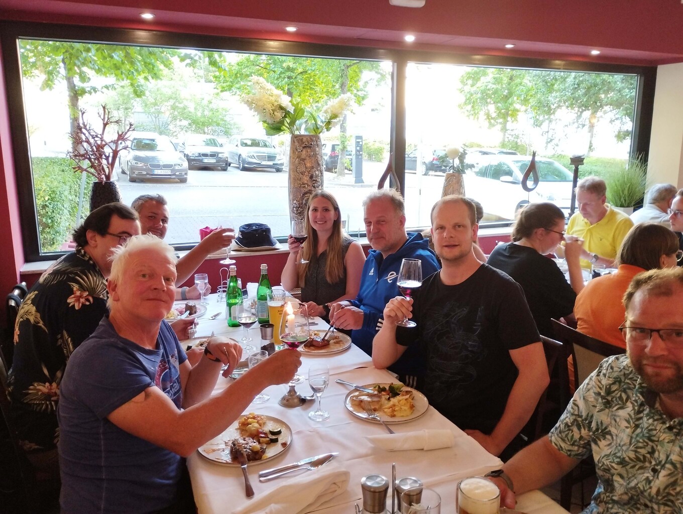 lachende Gruppe von etwa 8 Personen, die an einem Tisch in einem Restaurant sitzen und sich mit erhobenen Gläsern zuprosten