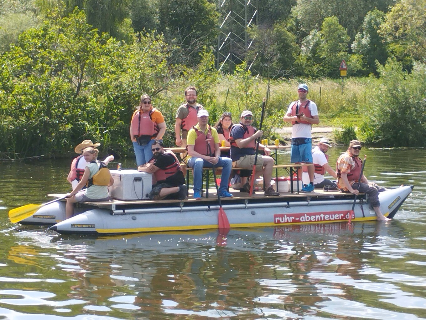 Gruppe von etwa 10 Personen, die auf einem selbstgebauten Floß mit zwei Pontons auf einem Gewässer unterwegs sind, die meisten tragen orangefarbene oder rote Schwimmwesten 
