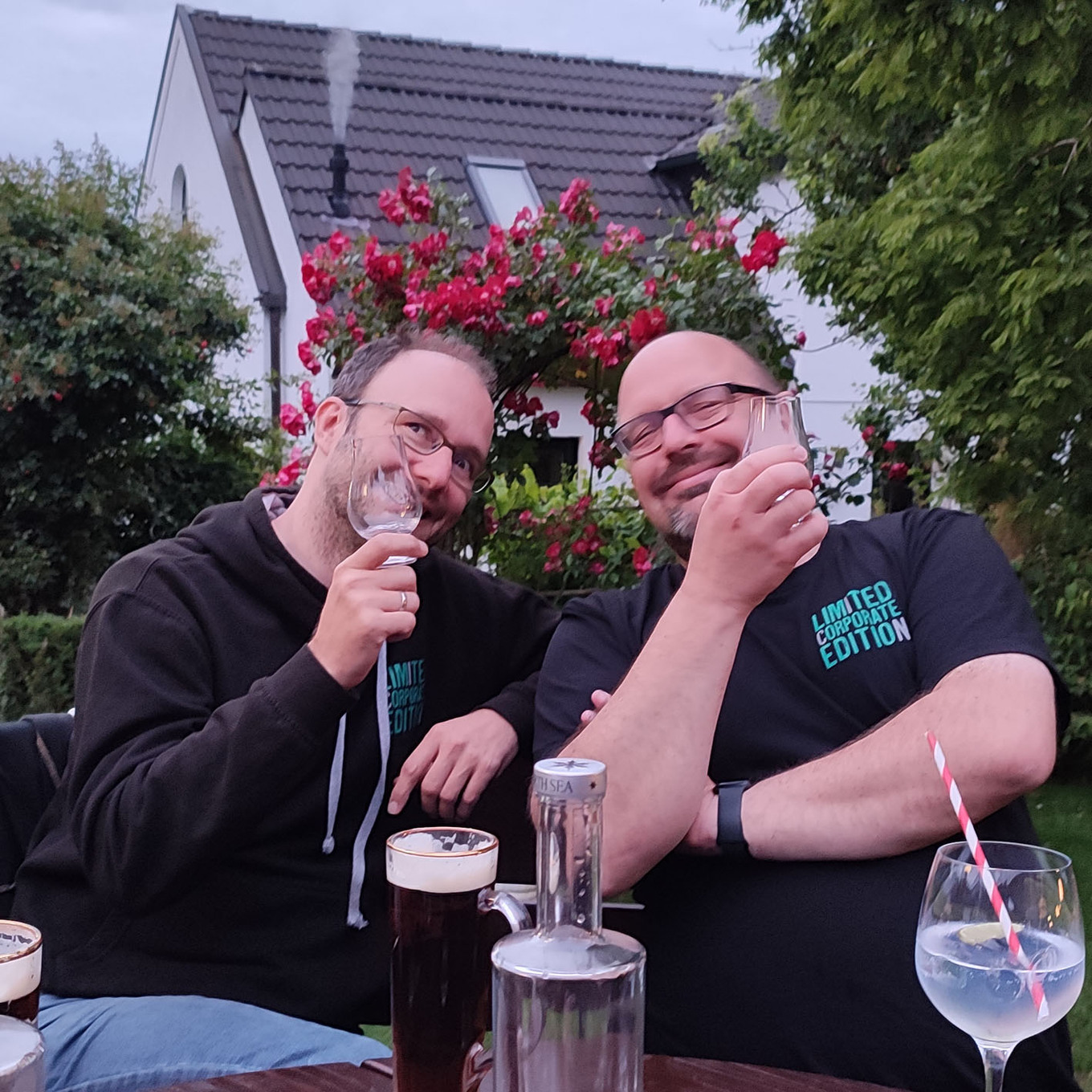 zwei lächelnde Männer, die im freien auf einer Terrasse sitzen und Gläser mit Getränken in der Hand halten 