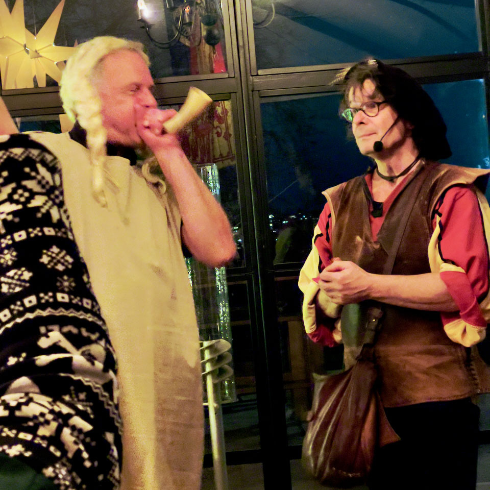 ein Mann in einer Verkleidung, der in ein großes Horn bläst, neben ihm steht eine andere Person in einer Ähnlichen Aufmachung, die zuschaut