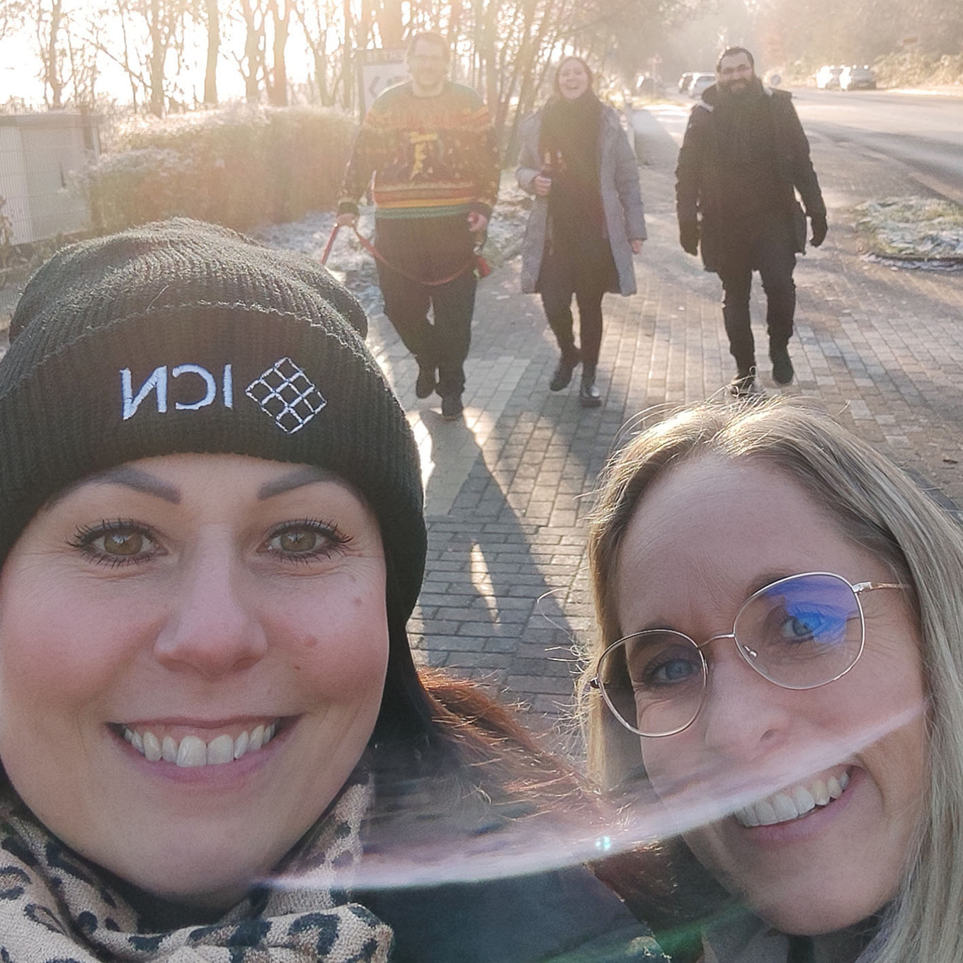 zwei Frauen in Nahaufnahme auf einem Selfie lächeln in die Kamera, im Hintergrund sind vier unscharfe Personen 