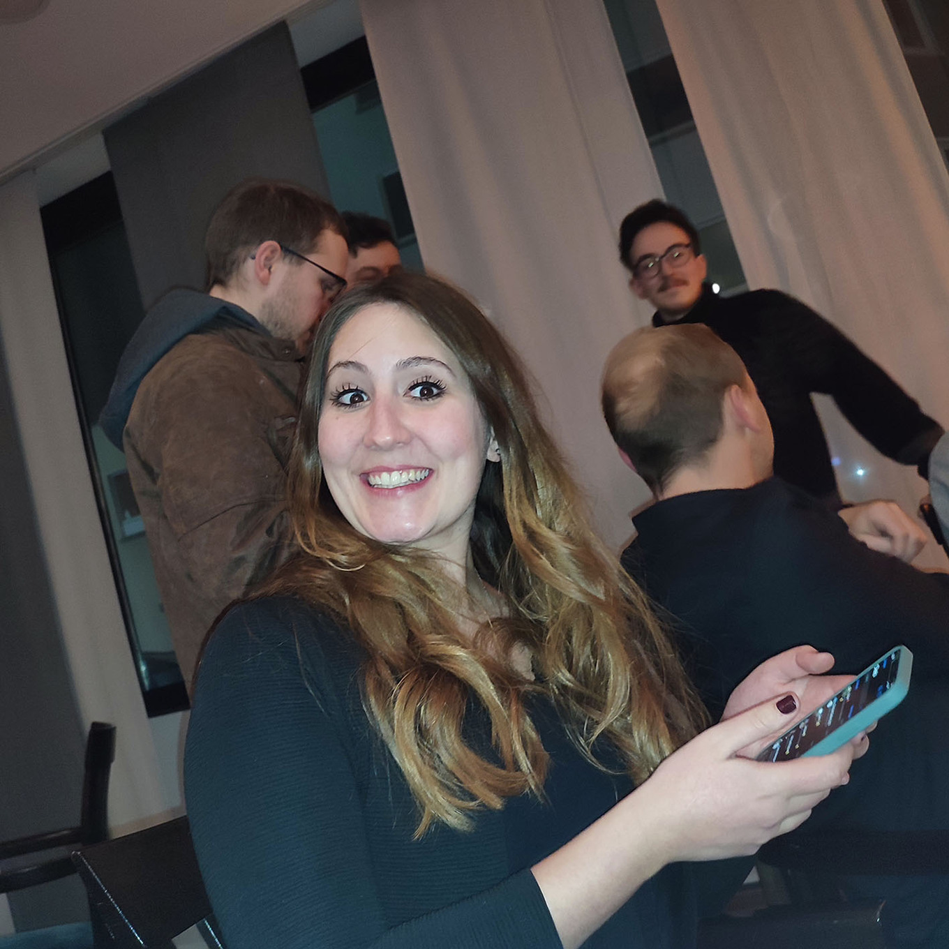 lachende Frau, die in die Kamera schaut und ein Smartphone in der Hand hält, im Hintergrund stehen mehrere Männer