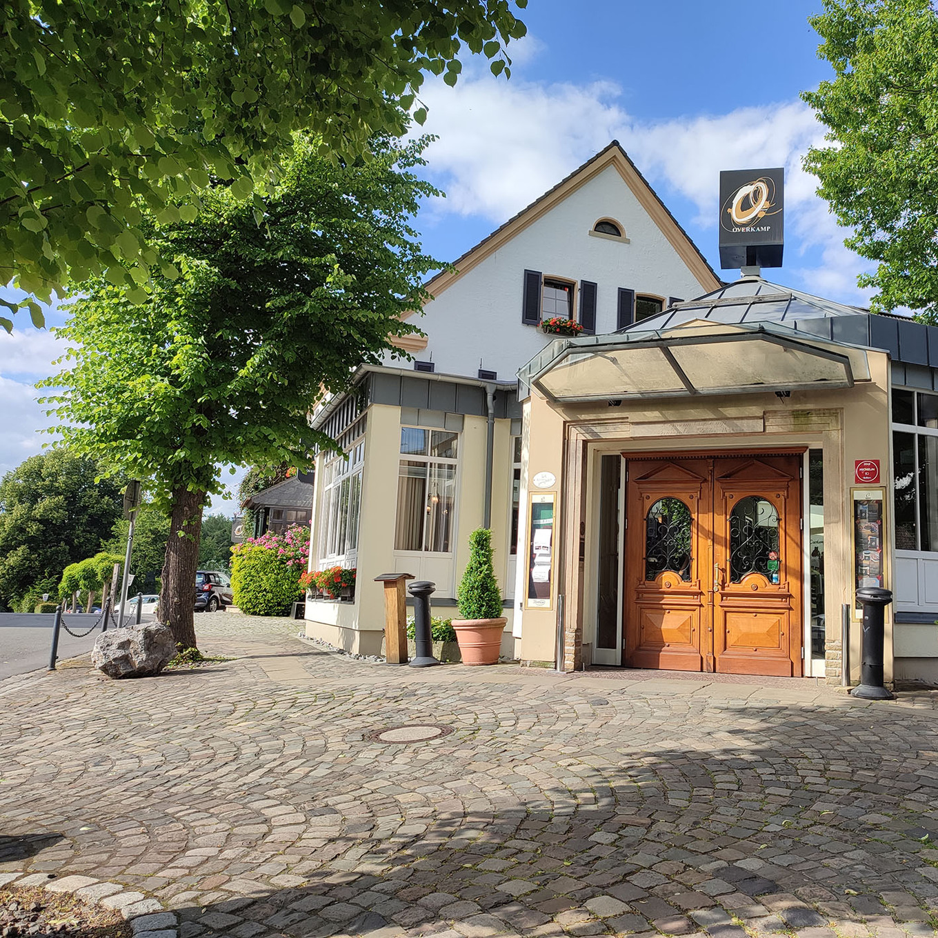 helles, zweistöckiges Gebäude mit einem Vordach aus Stein, umgeben von Bäumen, das eine hölzerne Doppeltür und ein Schild mit einem Firmenlogo hat