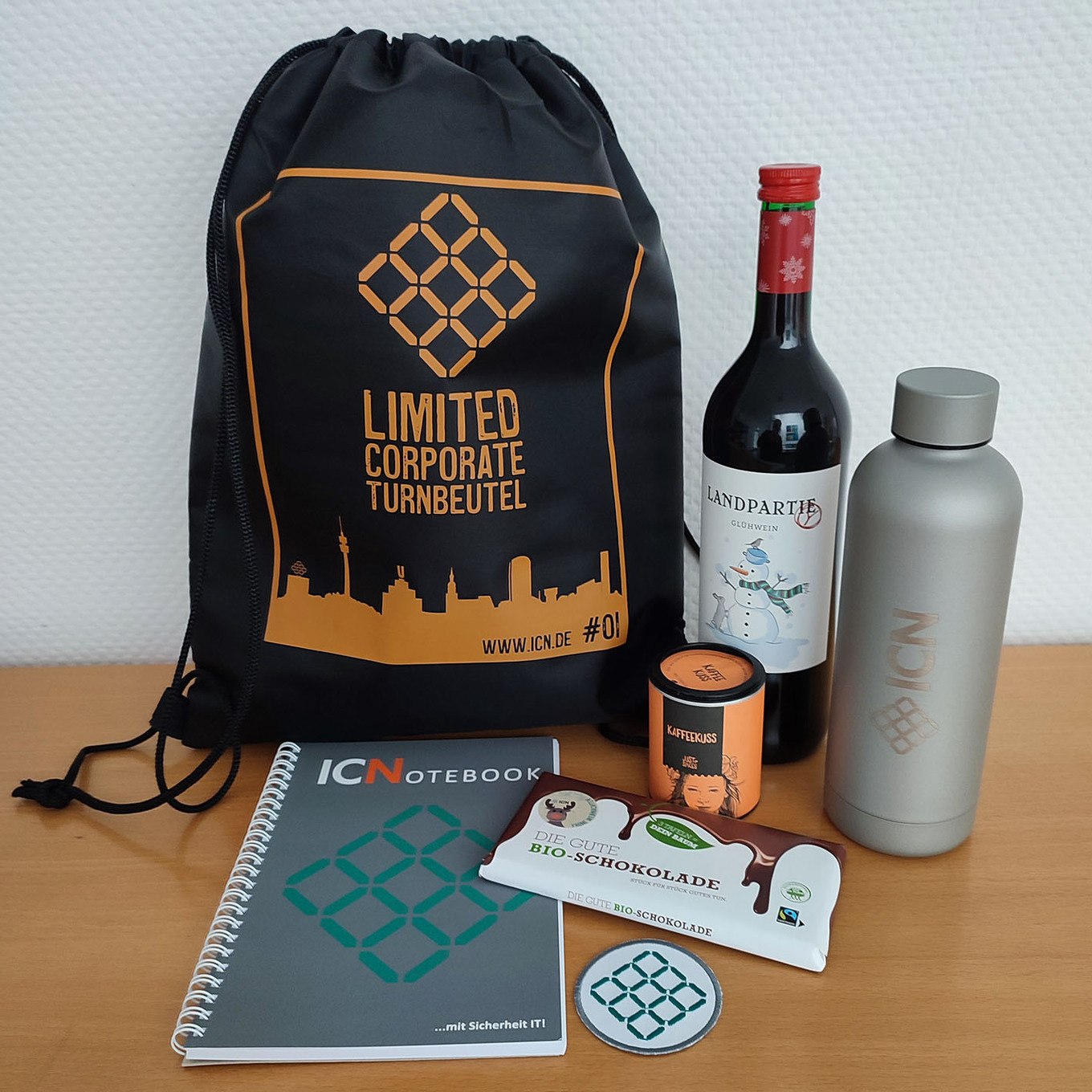 schwarze Sporttasche mit ICN Logo und der Aufschrift "United for Health", daneben eine Flasche Rotwein, eine silberne Trinkflasche, ein Notizbuch, eine Kerze und eine kleine Glasplatte mit ICN Logo 